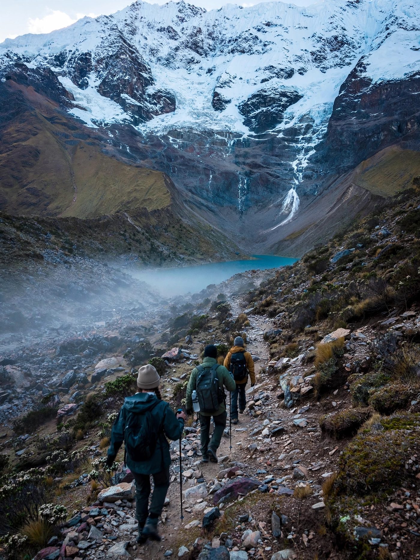 Viandanti in cammino verso una vetta andina innevata, tra nebbia e lago glaciale